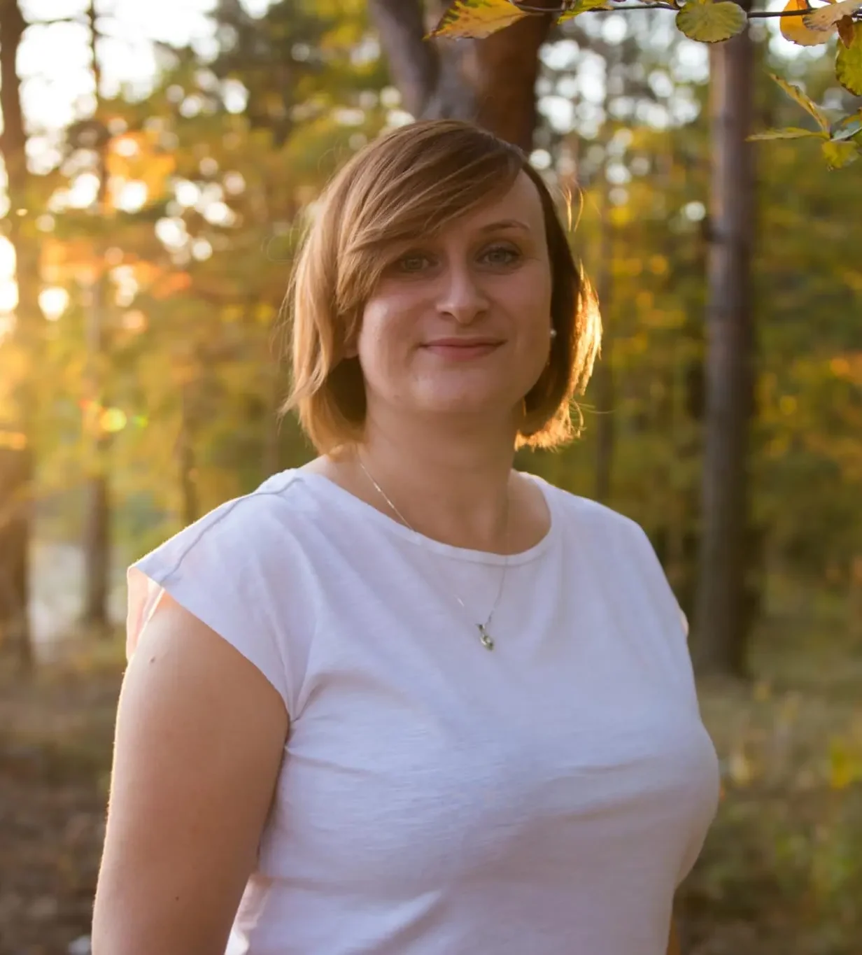 Kobieta w białej koszulce stojąca w lesie o zachodzie słońca, uśmiechająca się do aparatu.