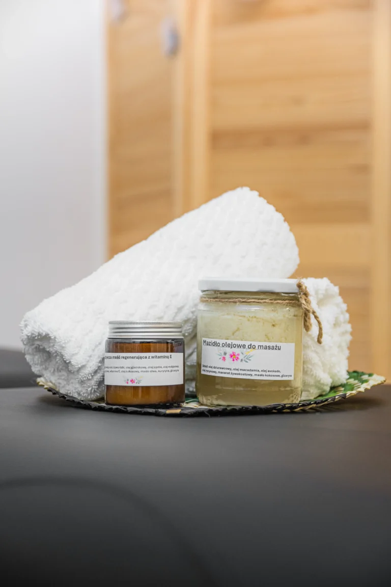 Naturalne kosmetyki do masażu w szklanych słoikach, biała ręcznikowa rolka i pleciona podkładka na czarnym stole w gabinecie spa.