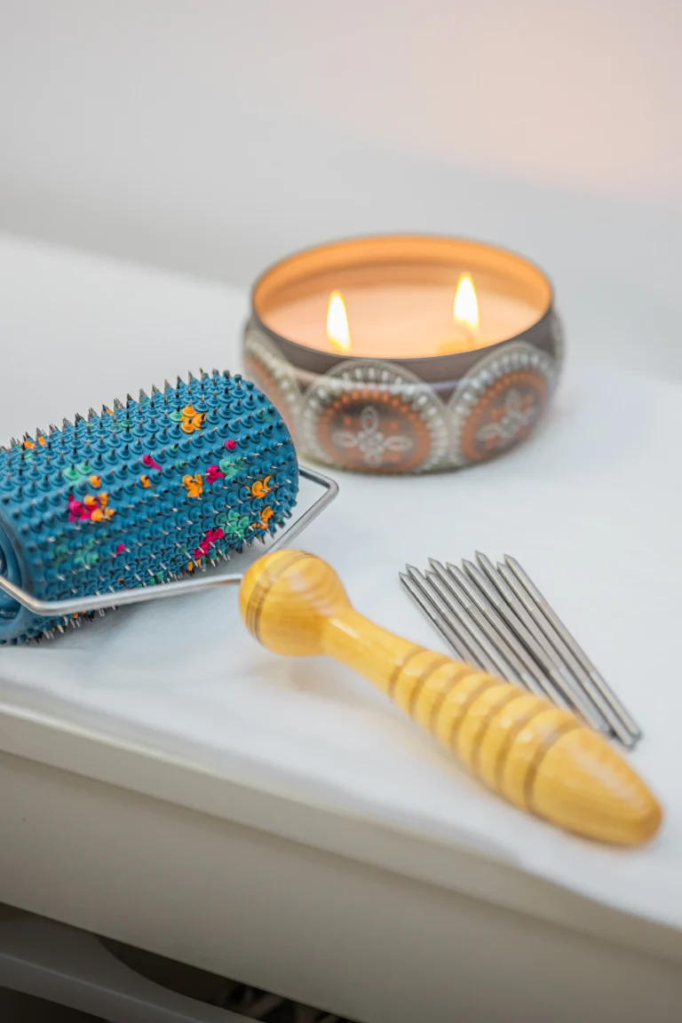 Kolczasty wałek akupresurowy, drewniany uchwyt, metalowe igły i zapalona świeca dekoracyjna na białym stole.