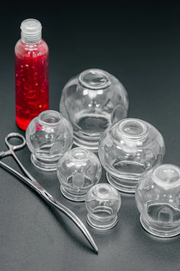 Zestaw szklanych baniek do terapii próżniowej, metalowe szczypce i butelka z czerwonym płynem na czarnym tle.