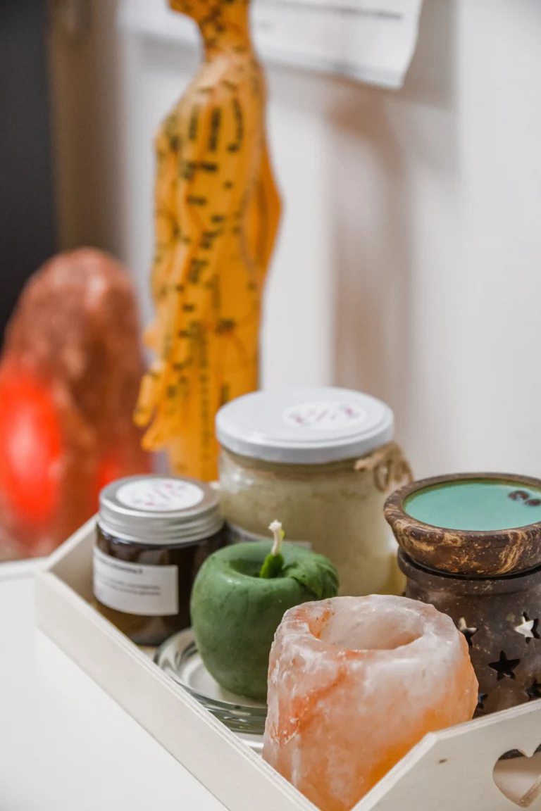 Zestaw naturalnych produktów na drewnianej tacy, w tym świece, słoiki z kosmetykami, lampa solna i figurka do akupunktury w tle.