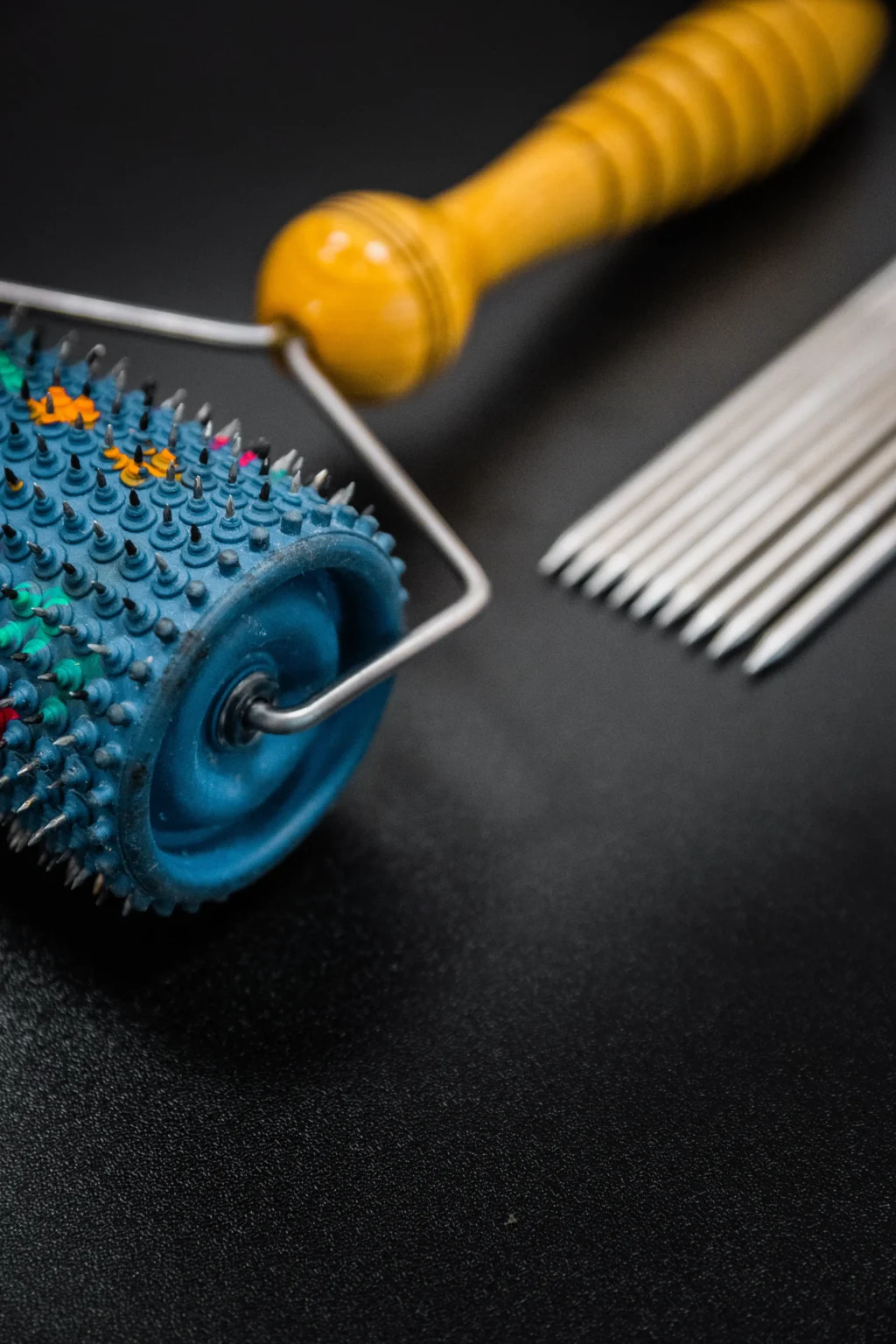 Kolczasty wałek do terapii akupresurowej i zestaw metalowych igieł na czarnym tle.